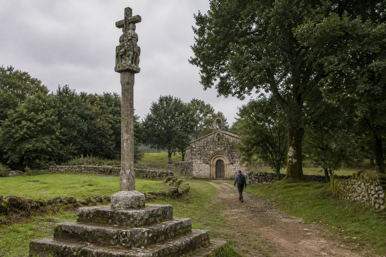 Cruceiro de granito junto a un sendero rural y una pequeña iglesia de tradición románica en el Camino